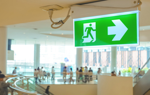 Green electric emergency exit sign on ceiling with blurred background of people at a shopping mall.