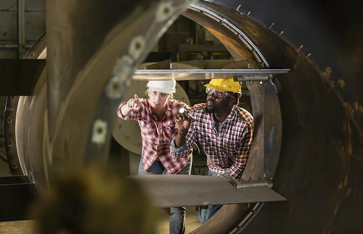 Two multi-ethnic workers in a metal fabrication factory inspecting a product with a flashlight.