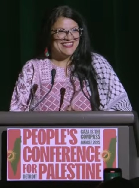 A picture of Rashida Tlaib standing at a podium with microphones. There is a sign on the podium that reads 'People's Conference for Palestine; Gaza is the Compass; Detroit; August 2025'.