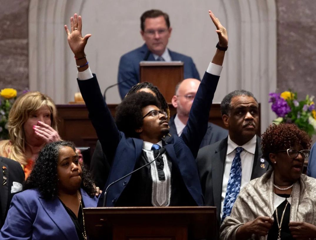 Justin standing at a podium in a suit with both of his arms raised above his head.