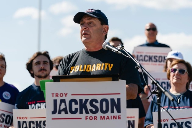 Photo of Troy Jackson speaking at an outdoor rally behind a podium, wearing a SOLIDARITY shirt, with supporters holding Troy Jackson signs behind him.