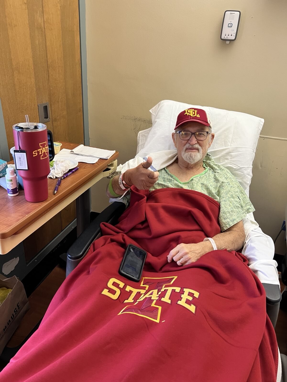 A picture of Katie Porter's dad laying in a hospital bed and giving a thumbs up towards the camera.
