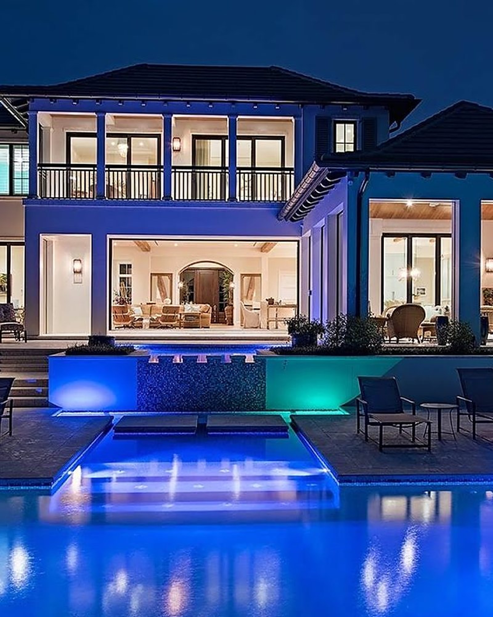Rear of a two-story estate at night, lit from within, with the pool in the foreground