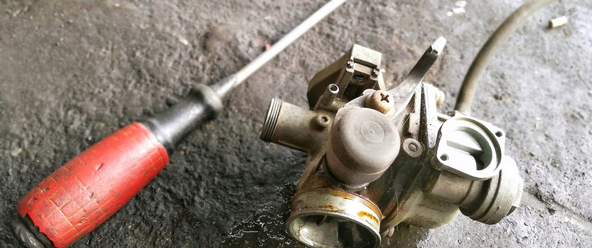 A used carburetor on a concrete floor next to a screwdriver.