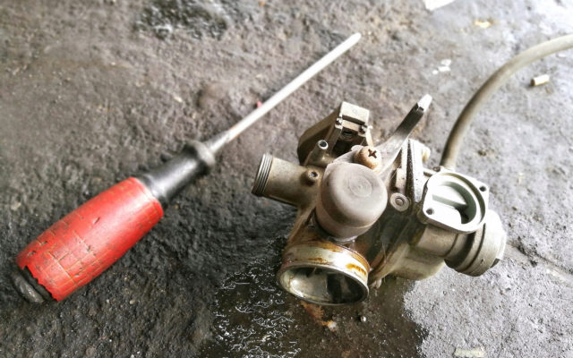 A used carburetor on a concrete floor next to a screwdriver.