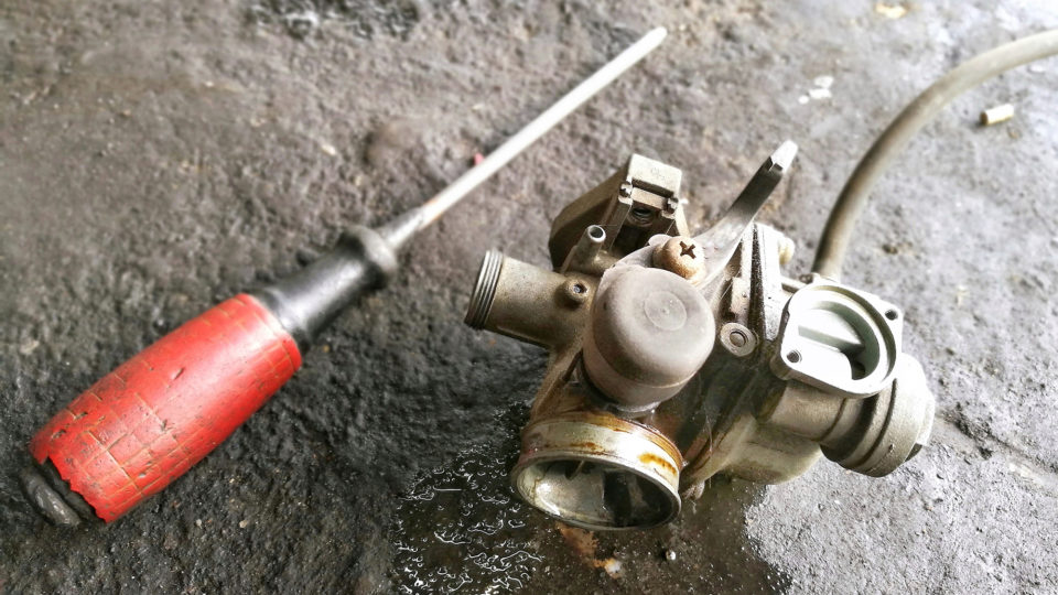 A used carburetor on a concrete floor next to a screwdriver.