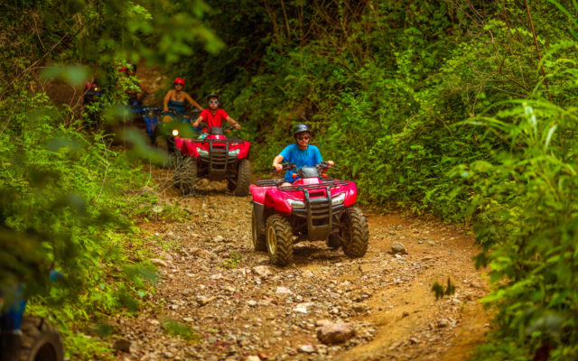A family rides ATVs down a dirt path through the woods.