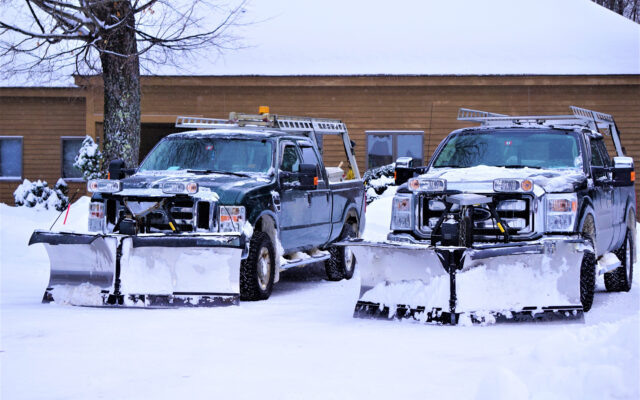 Two plows covered in snow in front of a building with snow on the ground.