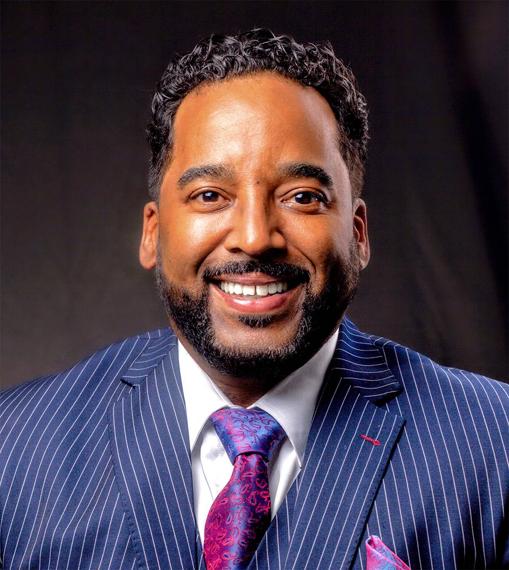 John Henry '95 smiling in blue pinstripe suit with colorful pink/purple tie and pocket square