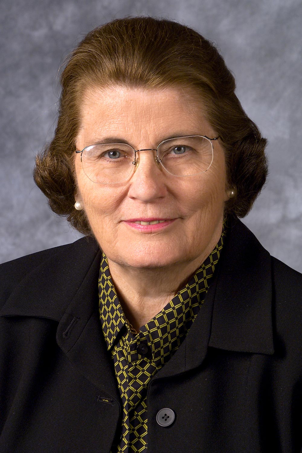 A portrait photograph of Virginia 'Ginny' Moriarty (P'79, P'86, P'94, G'11, G'19 - Long-standing supporter of Bucknell University) grinning in a black coat and black colored button-up dress shirt with a yellow x-crossed shaped style pattern on top of the dress shirt plus grey-tinted see through prescription glasses