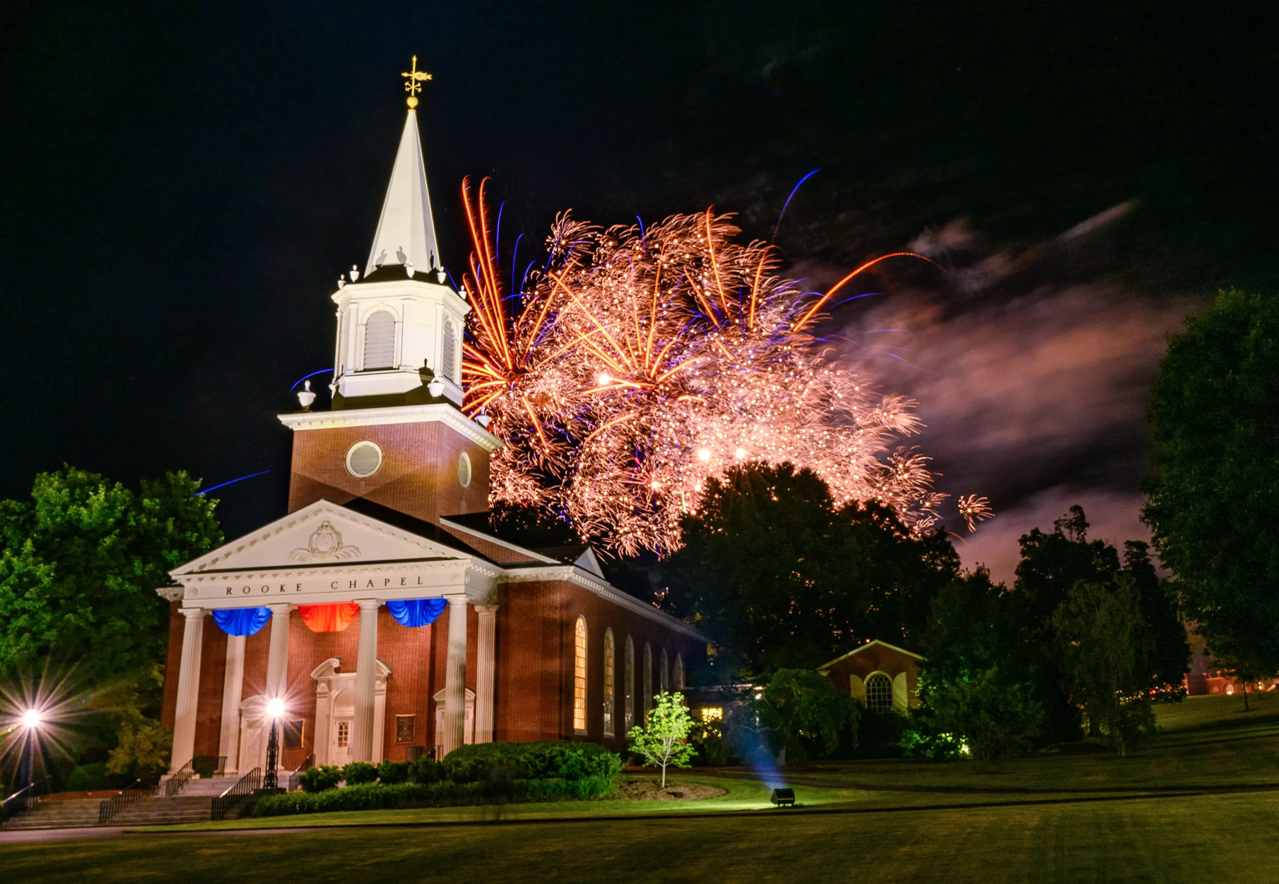 The sun breaks through trees that bower students relaxing in seasonally appropriate orange Adirondack chairs. Fireworks set off behind the Rooke Chapel for Reunion Week