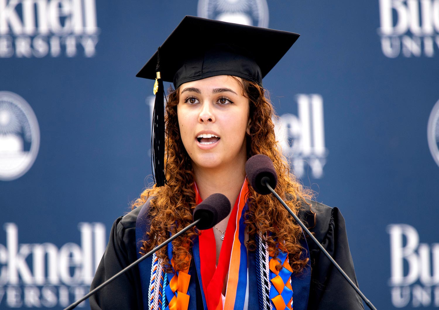 Kaia Rendo ’23 wearing a grad cap and gown speaks to the ceremony audience