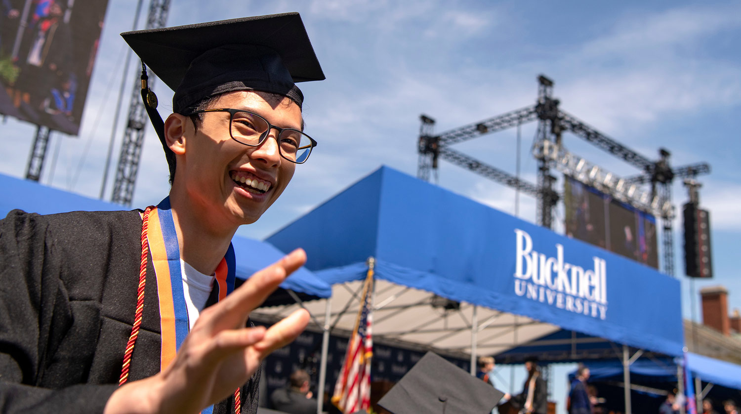 angled view of a grad smiling and waving with the ceremony stage visible in the distant background
