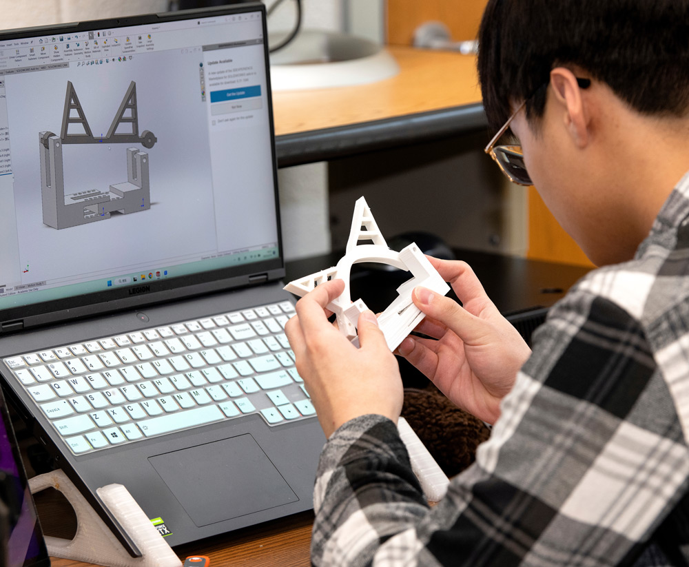 Student working on laptop and 3D printer holding piece in hand.