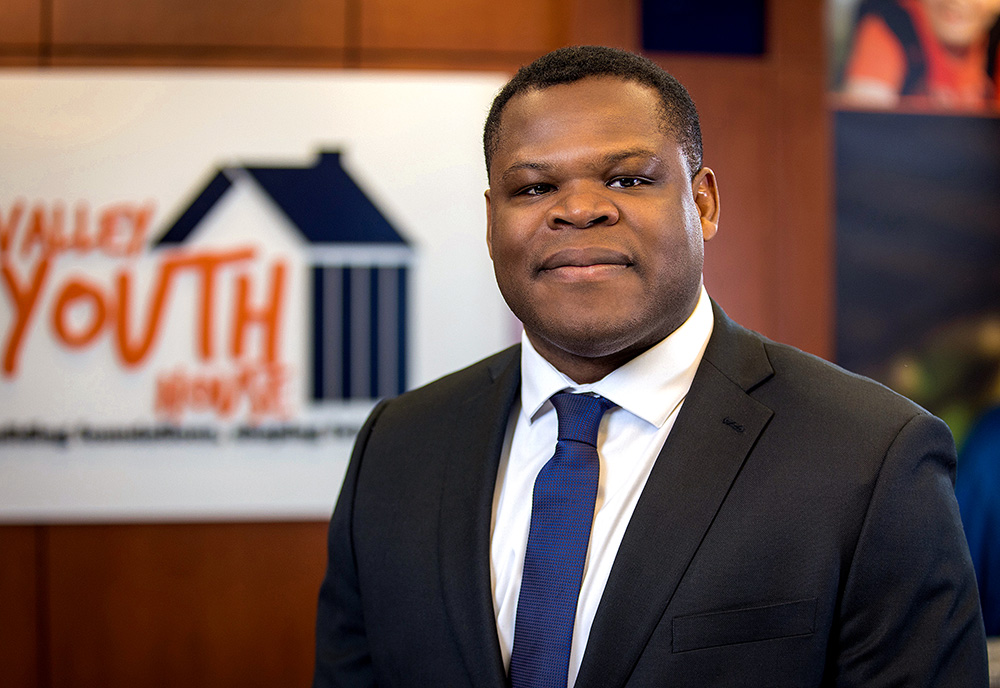 Portrait photograph of Tyler Greene '17 grinning in a black suit and white dress-shirt underneath plus dark blue tie inside the Valley Youth House building