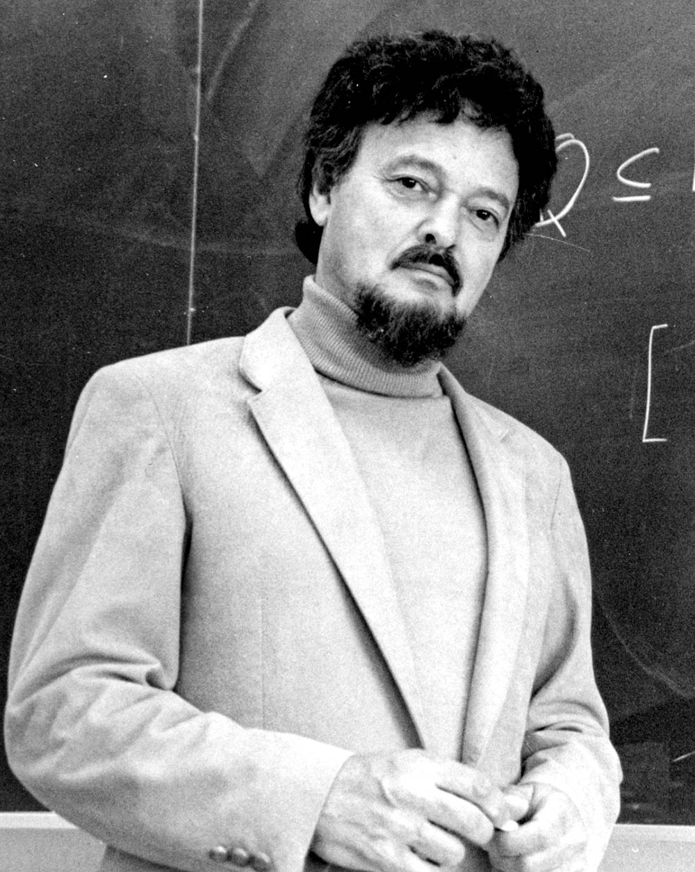 Close-up black and white portrait headshot photograph perspective of Charles Pinter P'87, P'94, P'02 posing for a picture in front of a chalkboard in a turtleneck and business coat open jacket while he plays with his fingers