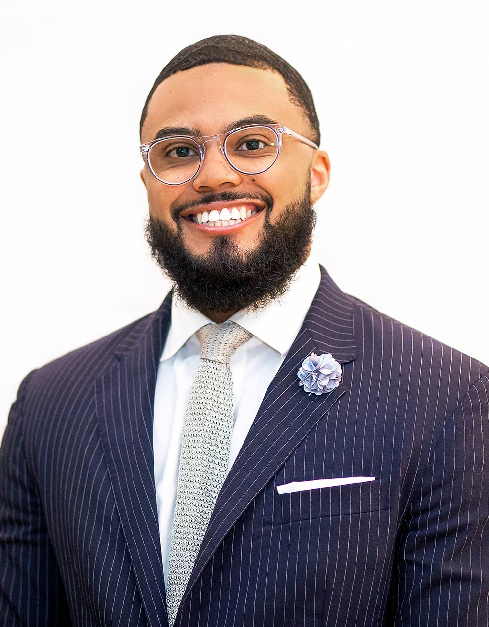 Close-up portrait headshot photograph perspective of Jaylen Amaker '15 smiling in a dark navy blue/white striped lines business suit and white button-up dress shirt with a pattern colored tie (faded gradient white/dark blue) plus little blue flower ribbon pinned on his left side of the suit