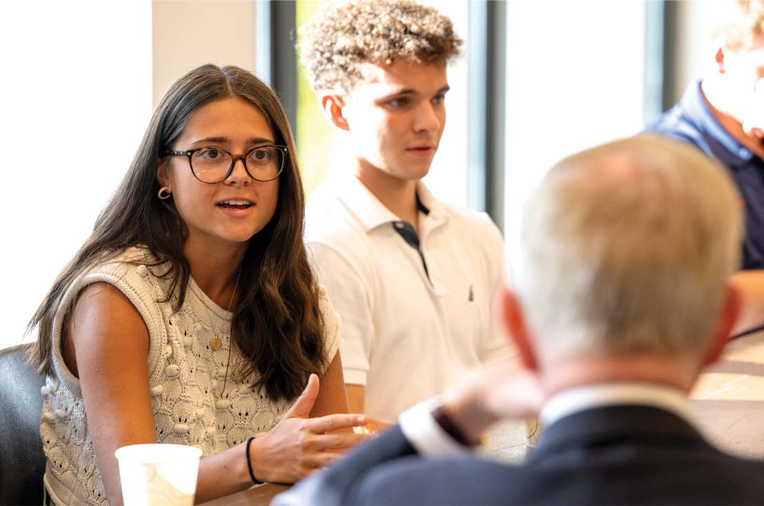 Young woman sitting at a table talking to a man who we see the back of his head