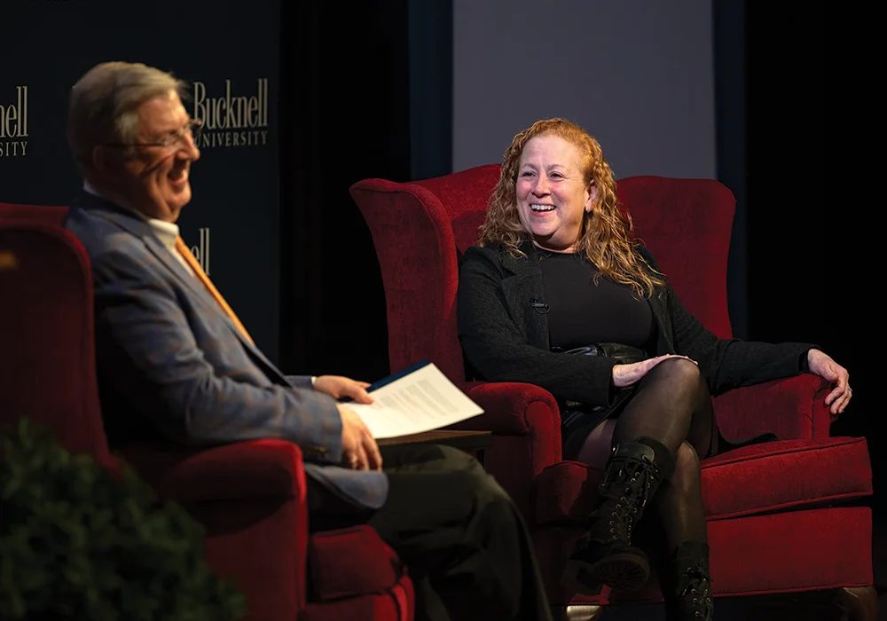 man sitting in red armchair wearing blue suit with yellow holding pieces of paper across from woman wearing all black out fit sitting in a red armchair smiling
