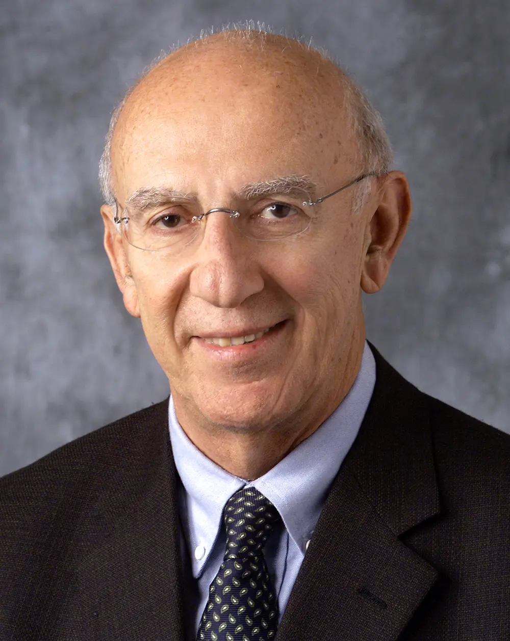 Portrait headshot photo of Edward Staiano smiling in a dark black business coat blazer open suit and light gray button-up closed dress shirt underneath equipped with a dark navy blue tie that has curved dark forest green pattern teardrop shapes all over