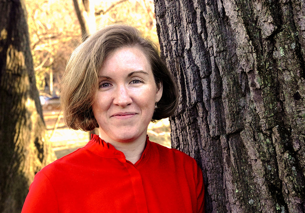 Portrait close-up headshot photo view of Alexandra Campbell-Ferrari in a bright red blouse standing/leaning up against the side of a tree trunk outdoors somewhere during the day