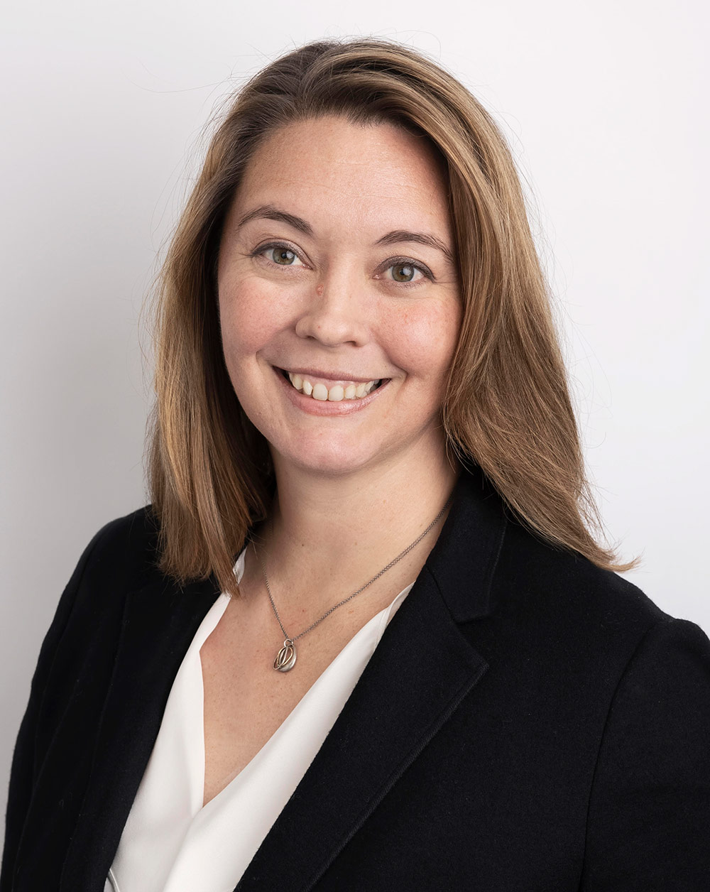 Portrait headshot close-up photo view of Michelle Lesh smiling in a black business coat blazer suit and white blouse underneath with a chrome-colored necklace around her