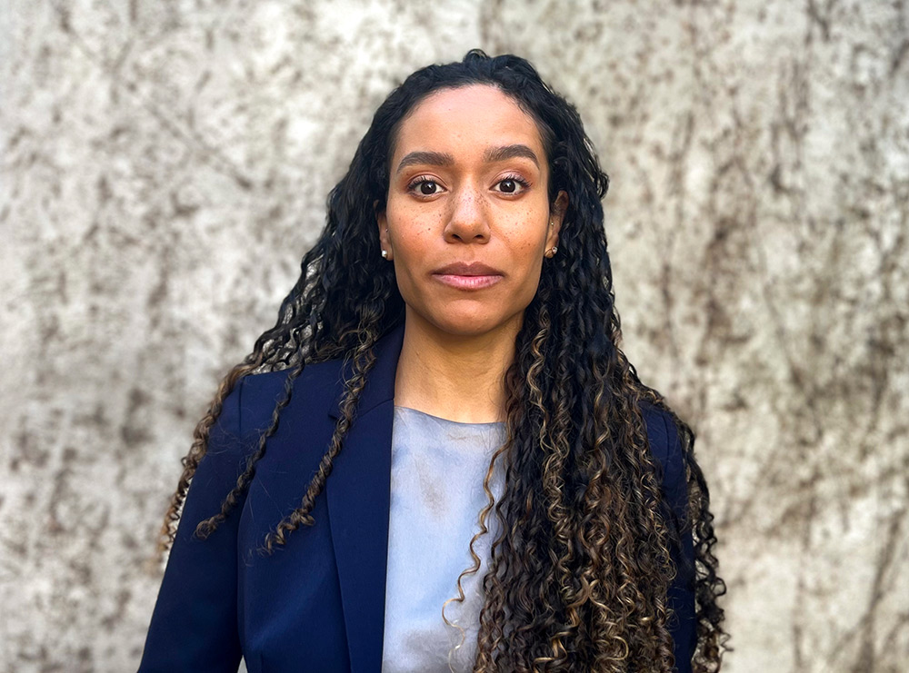 Portrait close-up photo view of Asha Jordan grinning in a dark navy blue business coat suit blazer and multi-chrome colored (light sky blue and dark grey) blouse underneath while she is standing outside somewhere
