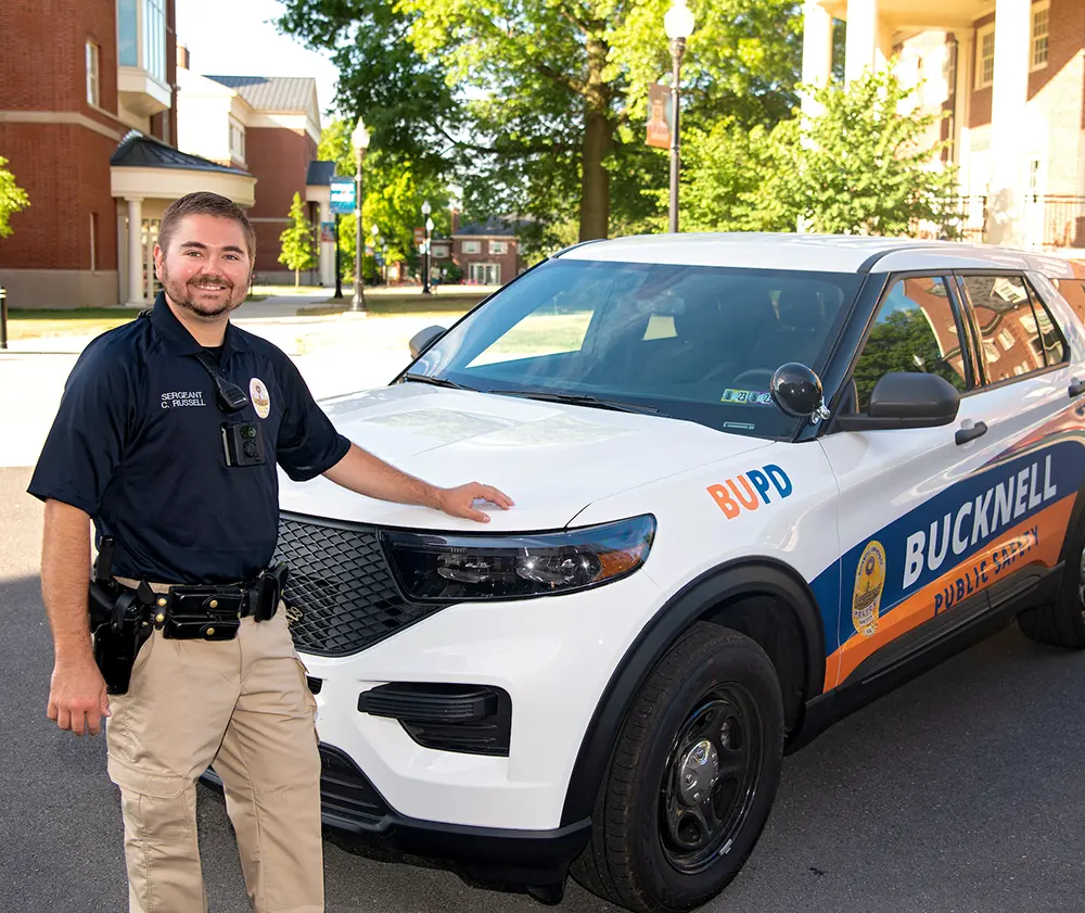 Portrait close-up photo view of Colbey Russell smiling in his Bucknell's Office of Public Safety officer uniform outfit featuring a dark navy blue polo top with his gold-colored badge equipped on it, his name imprinted in white on the polo top which says SERGEANT C. RUSSELL, and his other black-colored gadget work devices equipped on his polo top plus khaki light beige colored cargo pants as he poses with his left arm situated on top of the Bucknell Public Safety custom-themed SUV car on campus during the day