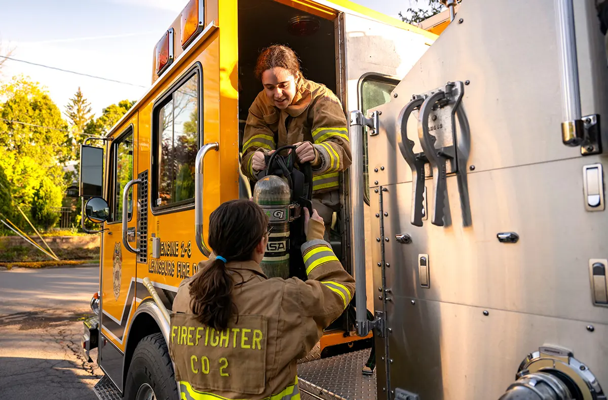 Danielle Kuck ’27 stands at a yellow fire truck, handing an oxygen tank to Isabel Byrnes ’27 who leans out from inside the truck