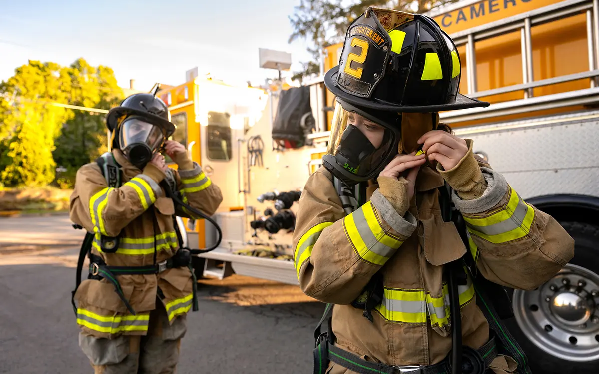 Danielle Kuck ’27 and Isabel Byrnes ’27 stand beside a yellow fire truck, adjusting the straps of their oxygen masks while in full bunker gear
