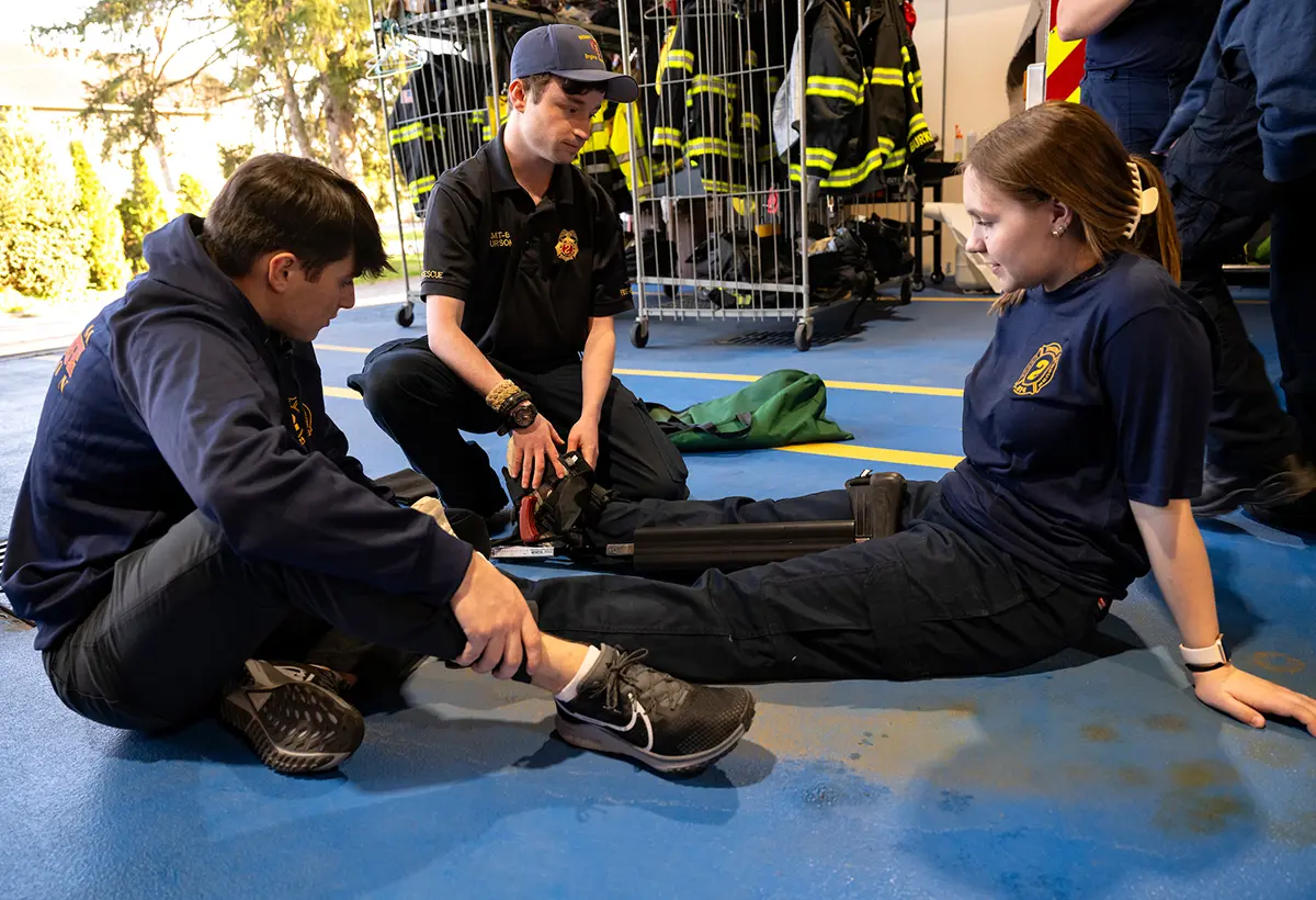 three students sit close on a fire station floor, one sits to the side as the mock patient while the other two students perform a leg splint exercise
