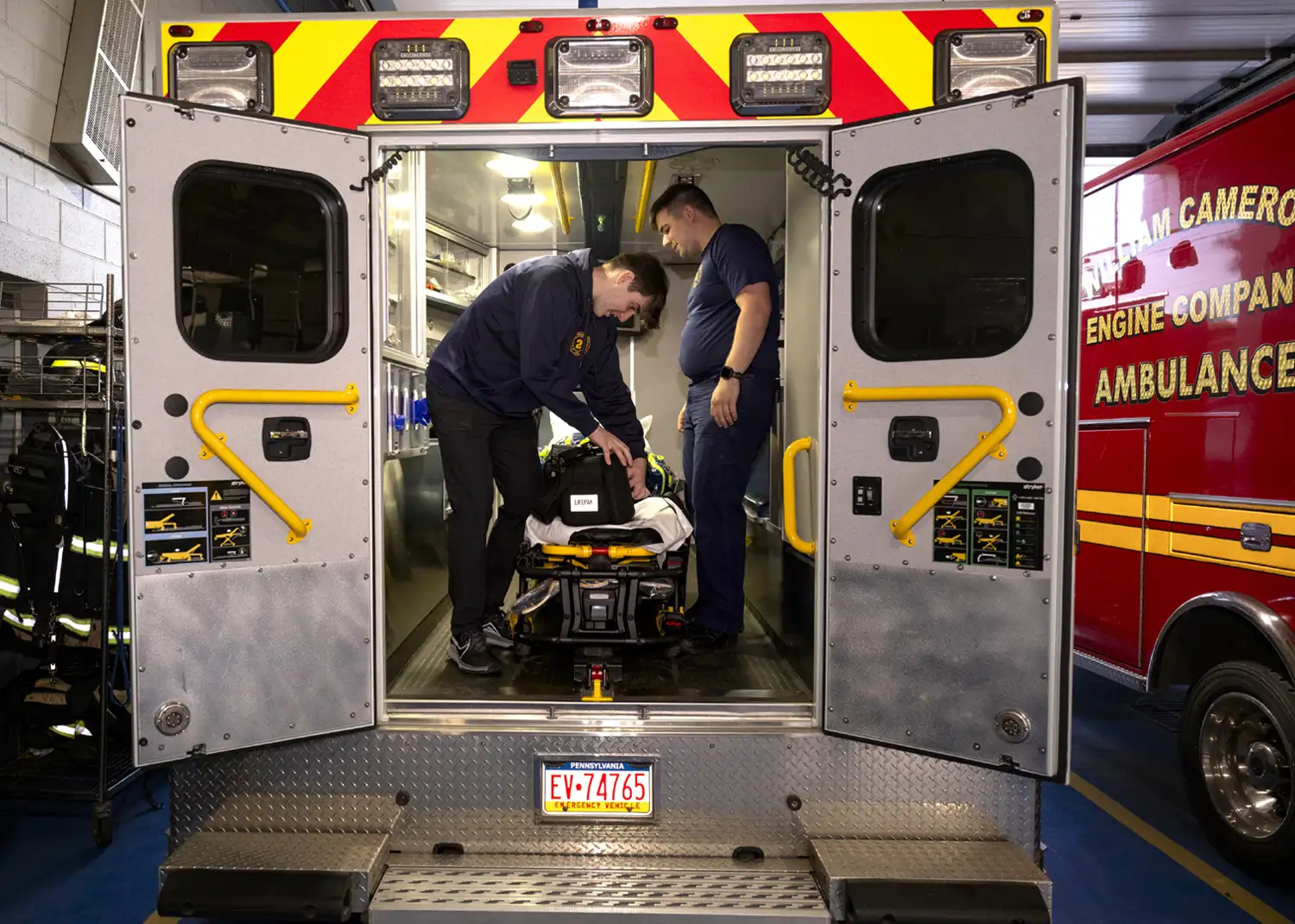two male students stand in the open back of an EMS vehicle parked in a fire station