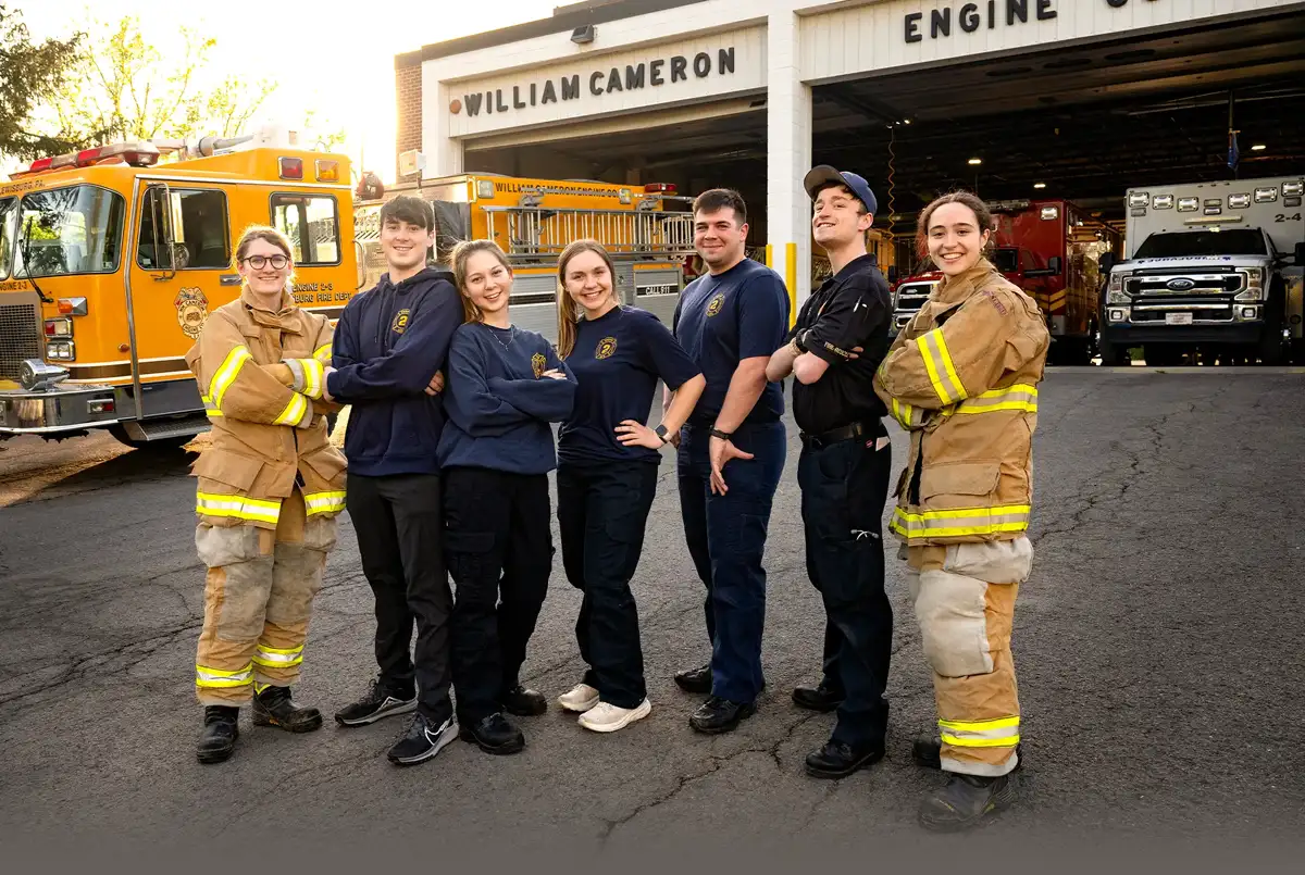 From left, Danielle Kuck ’27, Christopher Oko ’26, Kathryn Wrynn ’26, Olivia Jaye ’26, Matt Otto ’24, William Burson ’26 and Isabel Byrnes ’27 stand together smiling for a group photo in front of the William Cameron Engine Company with both bay doors open and a yellow fire truck and red and white EMT vehicles in the background