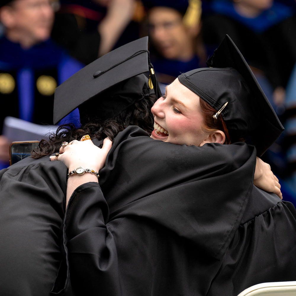 two students in cap and gown hugging at graduation ceremony