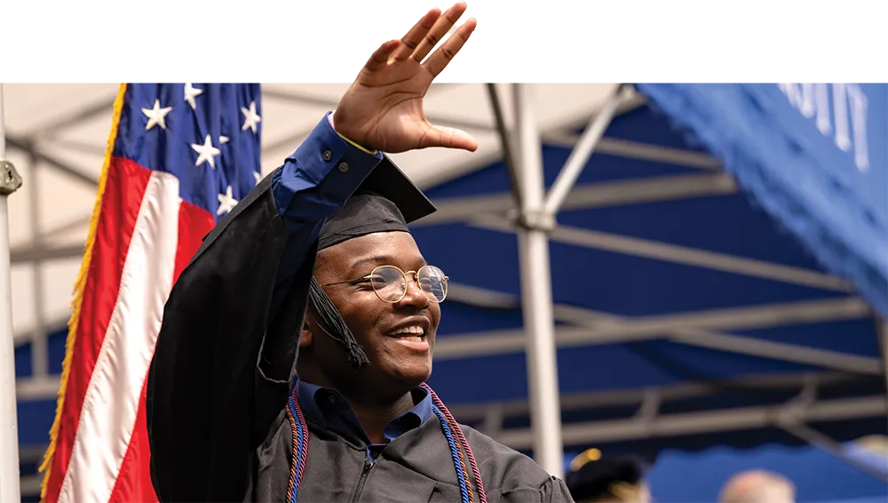student waving his hand on stage in cap and gown with an American flag behind him