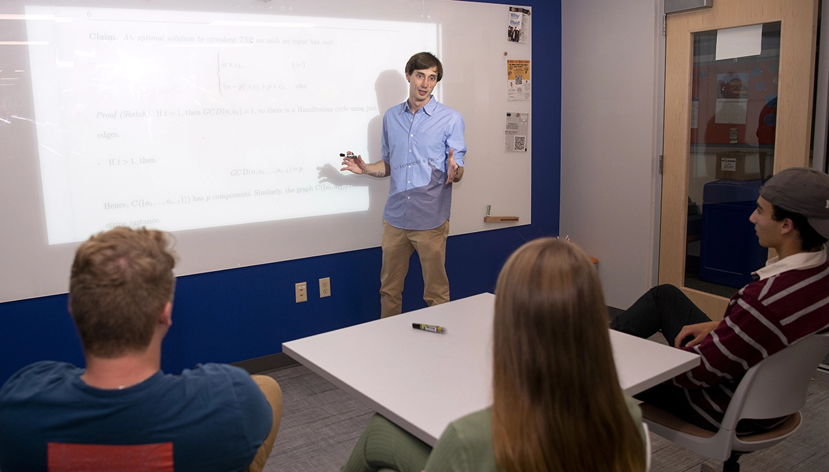 man holding marker speaking at front of classroom next to whiteboard with projection of math work and students sitting at desks watching