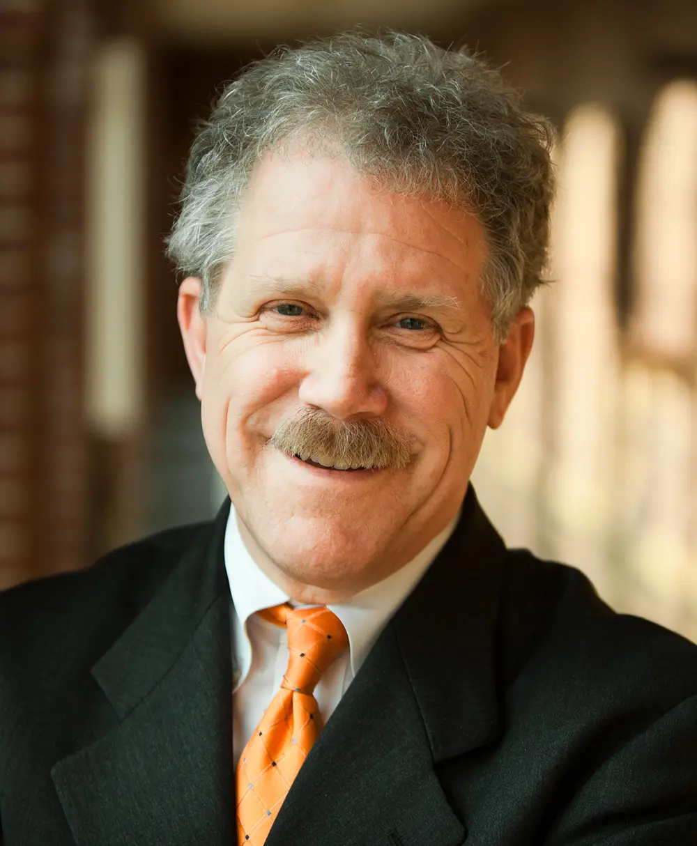 Portrait headshot close-up photo view of Michael Smyer smiling in a black business suit and white button-up dress shirt underneath and a orange gradient colored tie equipped in which the tie has tiny circular shapes and intersecting lines connecting throughout all these circular shapes