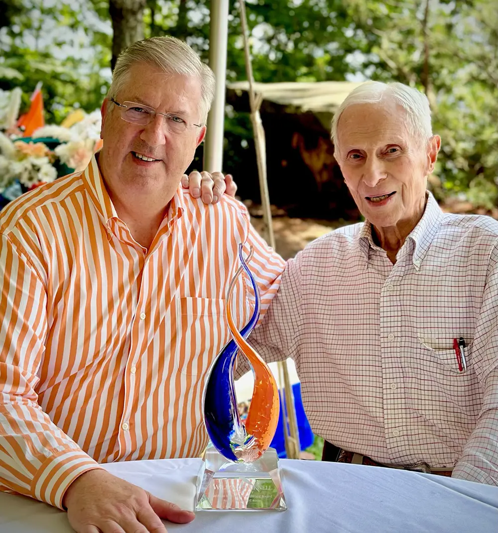 Portrait headshot close-up photo view of President John Bravman presenting Bob Rooke H’10, G’06, G’07, G’12 with the William Bucknell Philanthropic Award at a private event in July 2023 as both of them are smiling at a table seated down with the award situated on top of the table in the middle between them