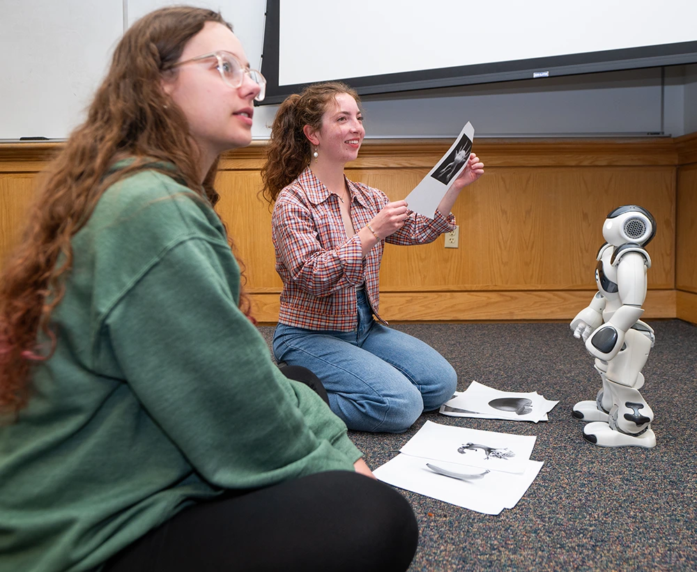 two female students seated on the ground with a small robot standing in front of them