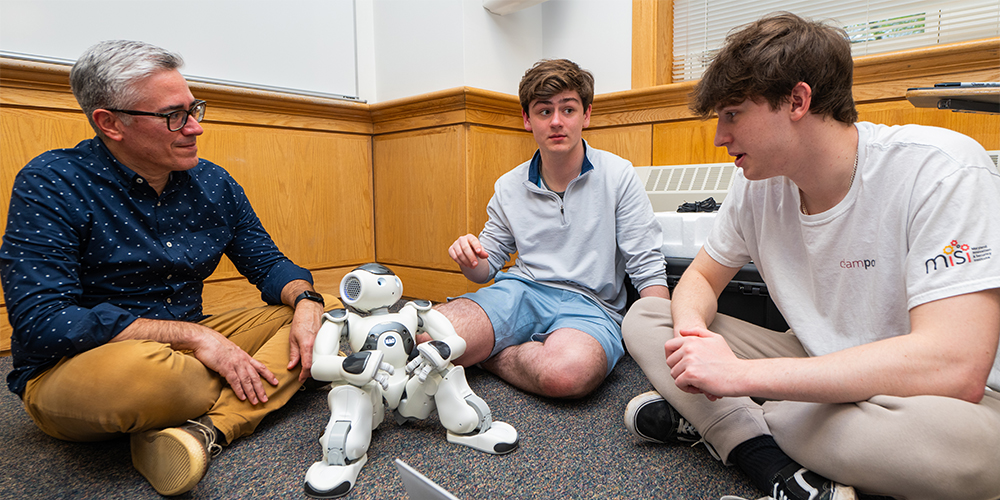 Professor and two students sitting on the floor with a laptop and small robot positioned in front of them