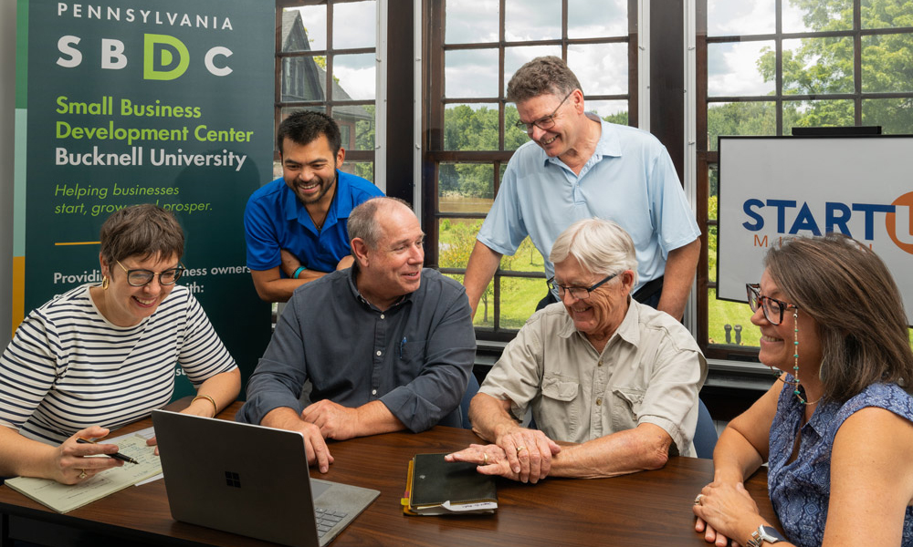 six people smiling and talking with gathered around a laptop