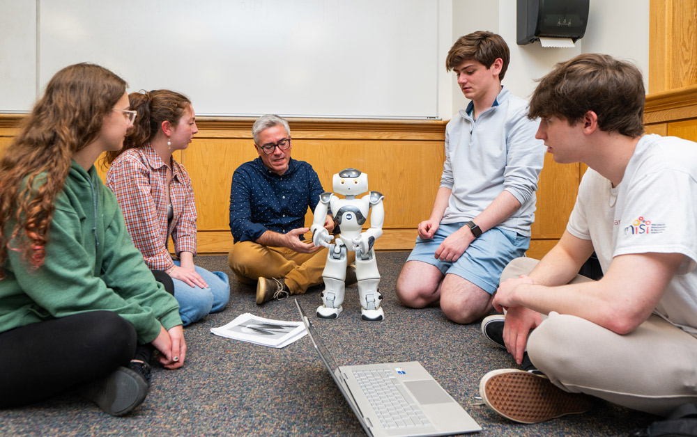 students and instructor seated on the floor while the instructor speaks and points at different parts on a robot