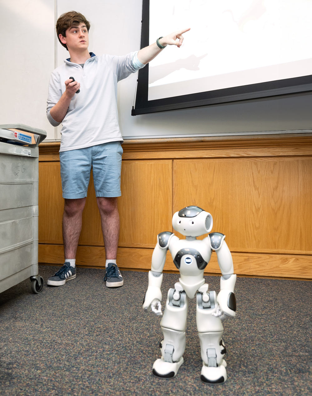 male student standing at the front of a classroom giving a presentation with a robot standing in front of him 