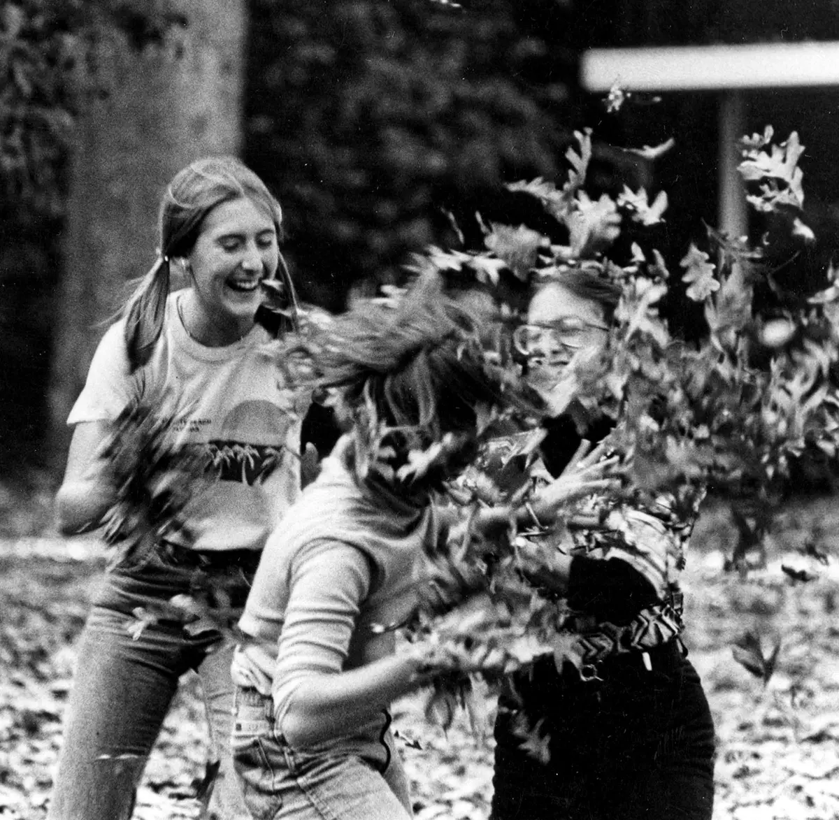 black and white photo of three Bucknell students throwing leaves at one another while laughing