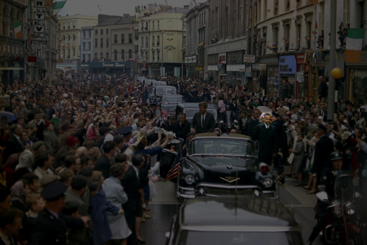 color photo of President Kennedy as he travels by motorcade through a crowd filled street in Cork, Ireland, with White House Secret Service agents Gerald A. "Jerry" Behn, Jerry Blaine, Bob Burke, Win Lawson, and Sam Sulliman close by