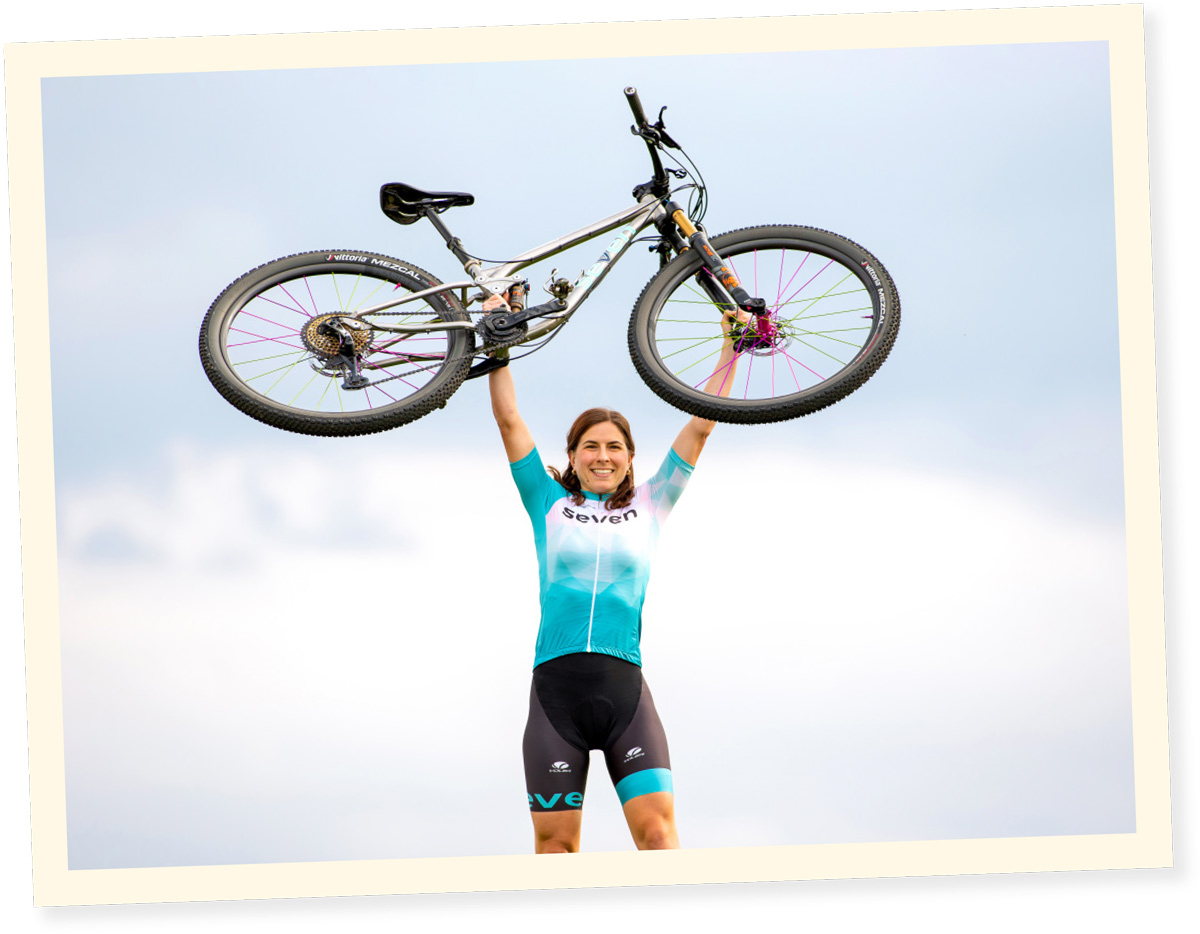 wide shot of Kelly Desharnais Catale hoisting her mountain bike overhead while wearing a wide smile