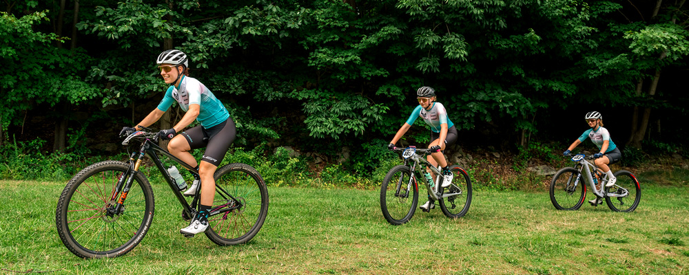 action shot of Kelly Desharnais Catale ‘12 leading KellCat Devo members Schuyler Hagge and Tessa Avery (L to R) on a biking trail