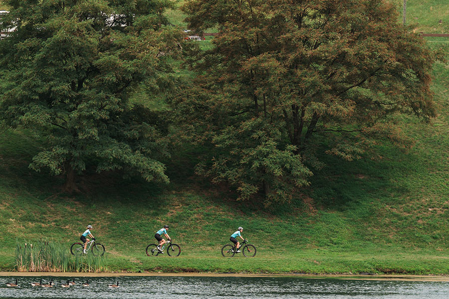three cyclists in matching jerseys stand with their bikes on grass, surrounded by trees