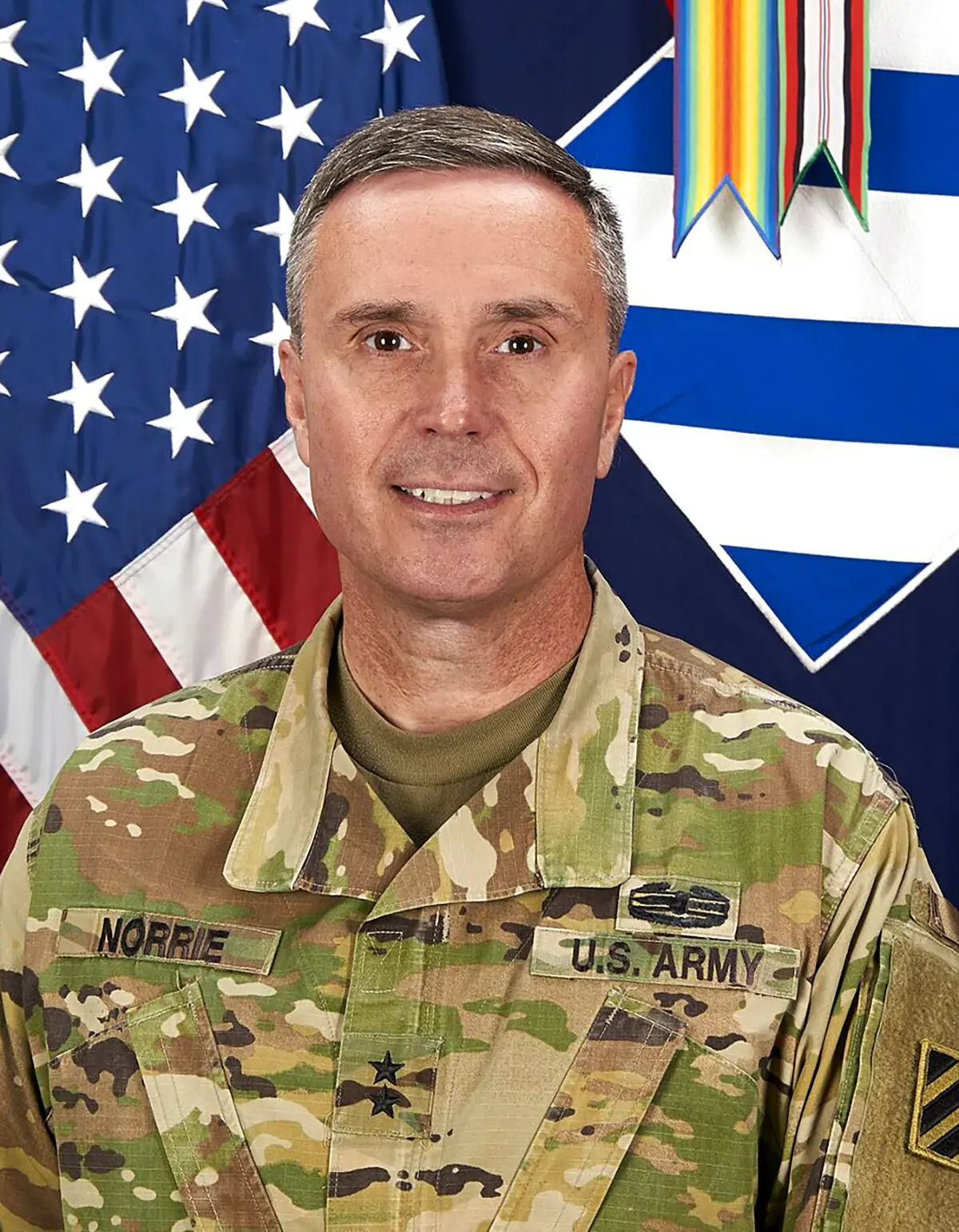 Portrait headshot photograph of Major General Chris Norrie smiling in his U.S. Army camouflage military attire outfit as he stands in front of a United States of America flag background