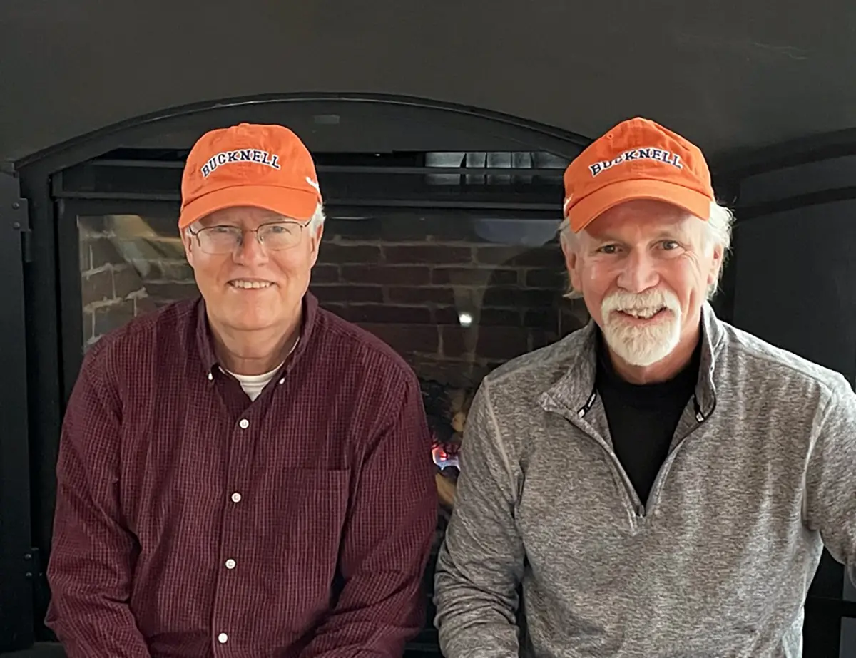 Portrait close-up indoor photo view of Richard J. Jones ’74, P’04 and T. Michael McCormick ’72, M’80 (pictured left to right) smiling as both of them have a orange Bucknell hat equipped as they are seated beside each other in front of a house chimney presumably